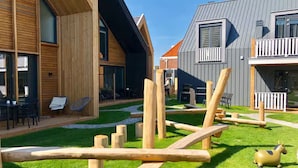 Un espacio exterior moderno con estructuras de juego de madera, césped y un edificio con ventanas amplias y un balcón.