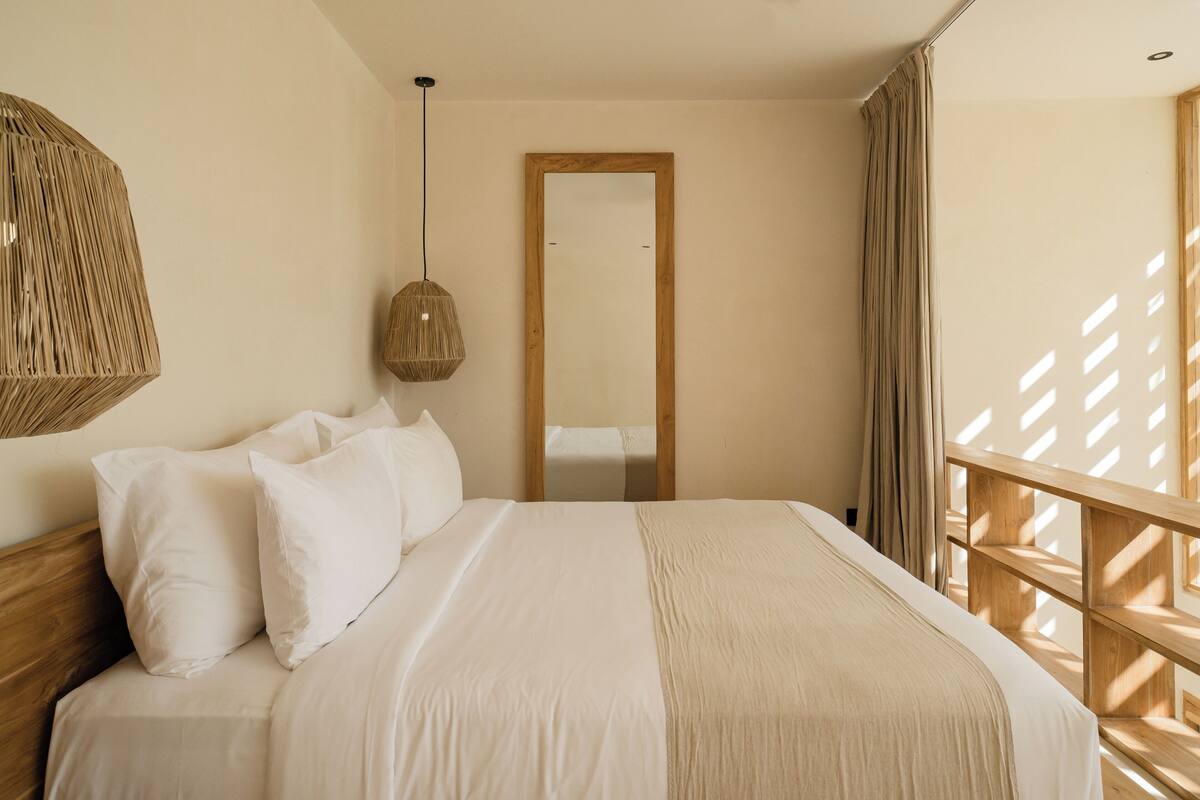 Ein Schlafzimmer mit Bett, Kissen, einer Pendelleuchte, einem Holzkopfteil, einem Spiegel und einem Fenster mit Vorhängen.
