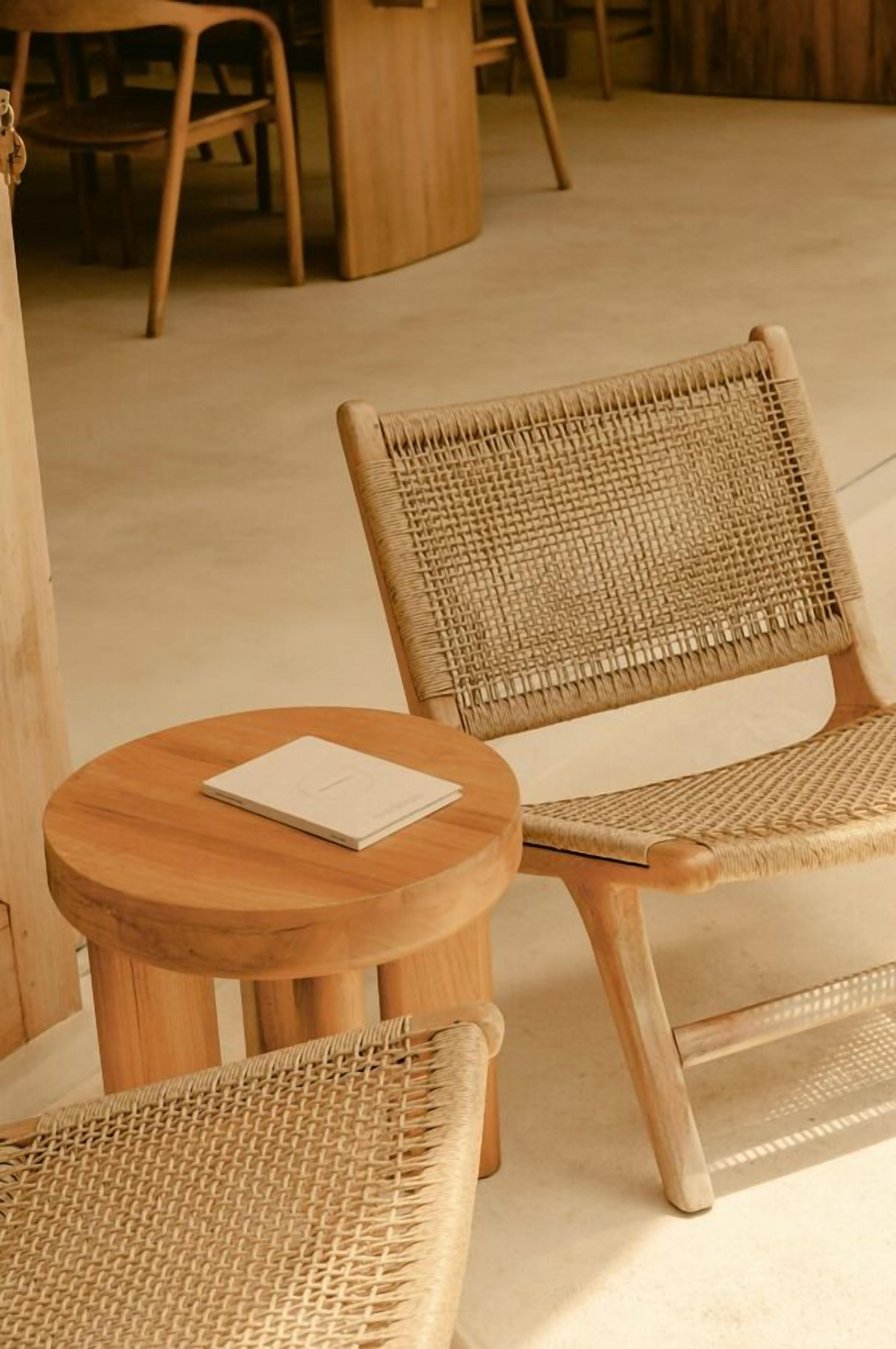 A wicker chair and a small wooden table with a book on it, in a room with a wooden floor and chairs in the background.