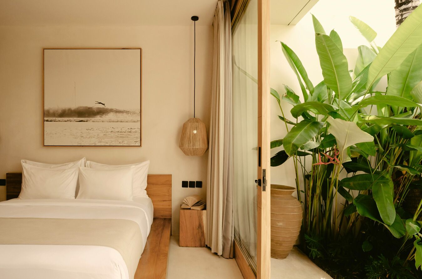 A bedroom with a bed, a bedside table with a book, a wall-mounted lamp, and a large window with curtains.