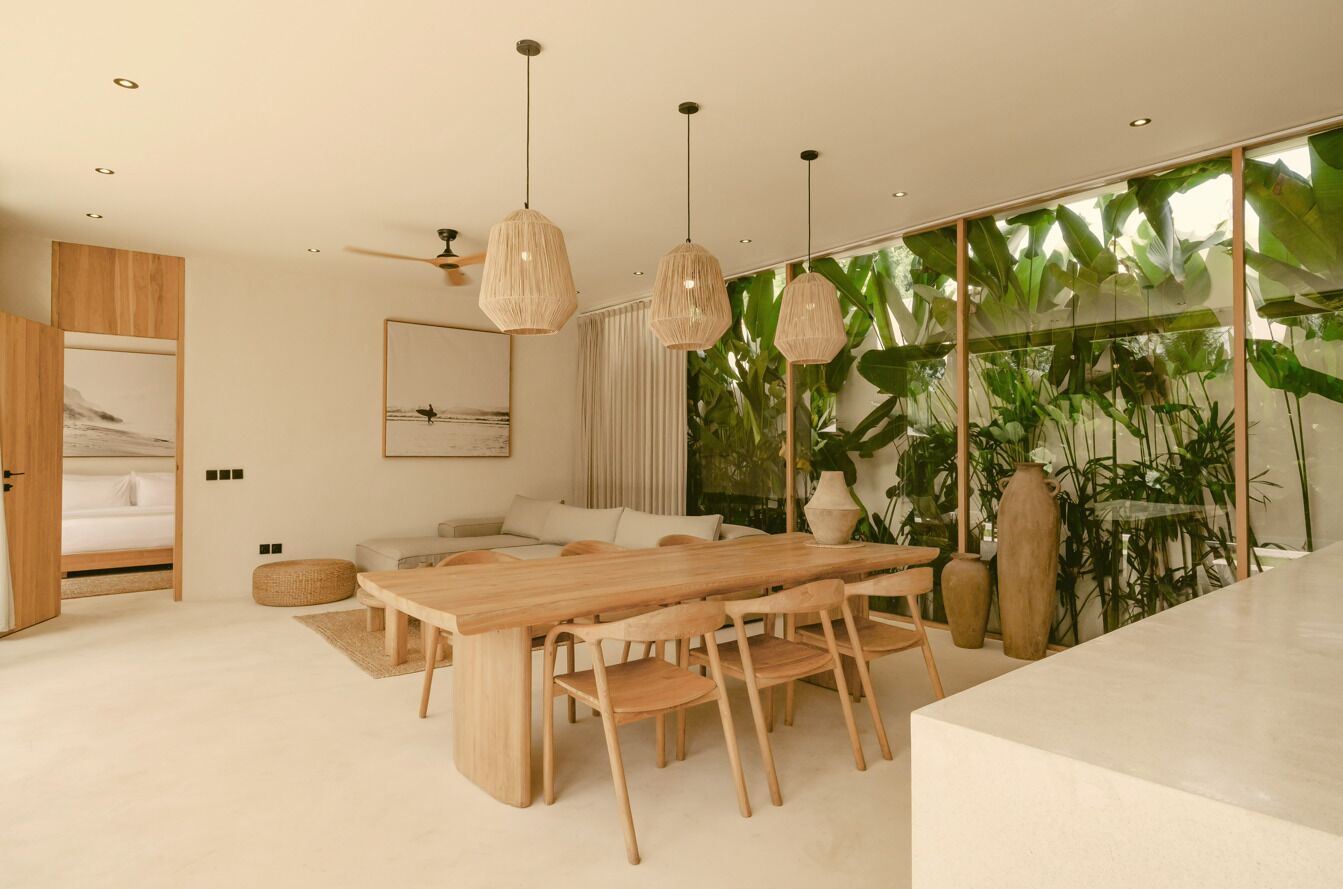 A modern living room with a wooden dining table, a sofa, and a large window with tropical plants.