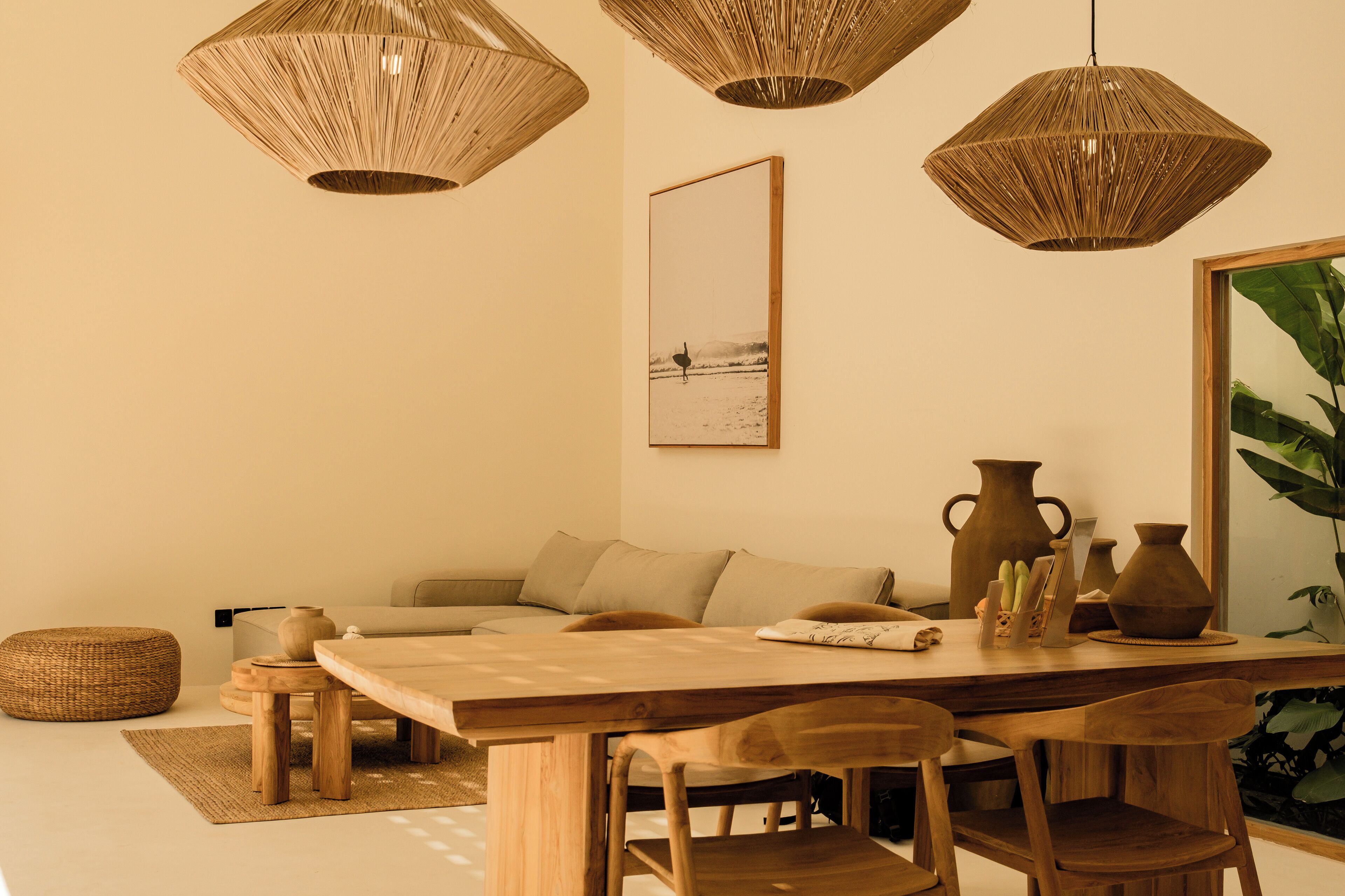 Un salon chaleureux avec une table à manger en bois, deux suspensions en fibres tressées, un canapé et un grand tableau encadré accroché au mur.