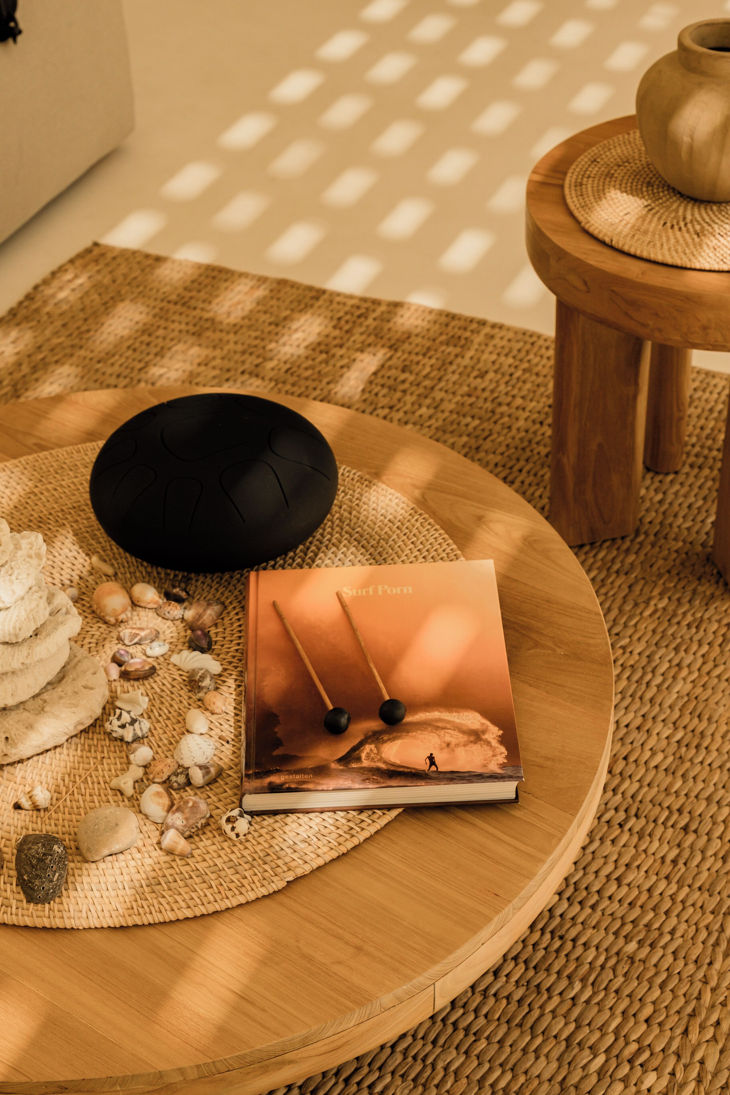Una mesa redonda de madera con un mantel tejido, una pelota de meditación negra, conchas marinas, un libro titulado 'Surf Poem' y un taburete de madera con una maceta.