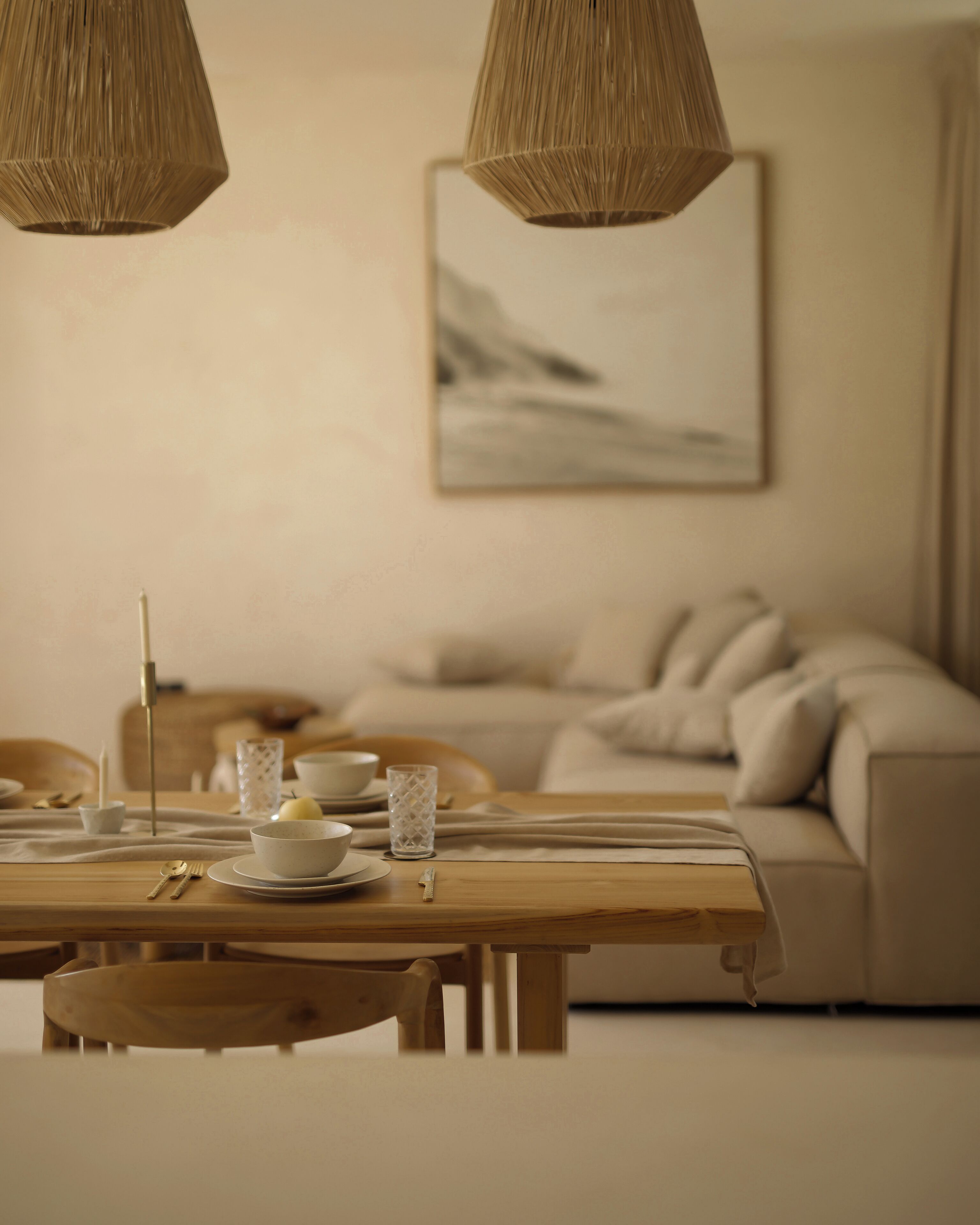 A dining area with a wooden table set for two, a candle, and a glass. A sofa and a painting are in the background.