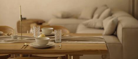 A dining area with a wooden table set for two, a candle, and a glass. A sofa and a painting are in the background.