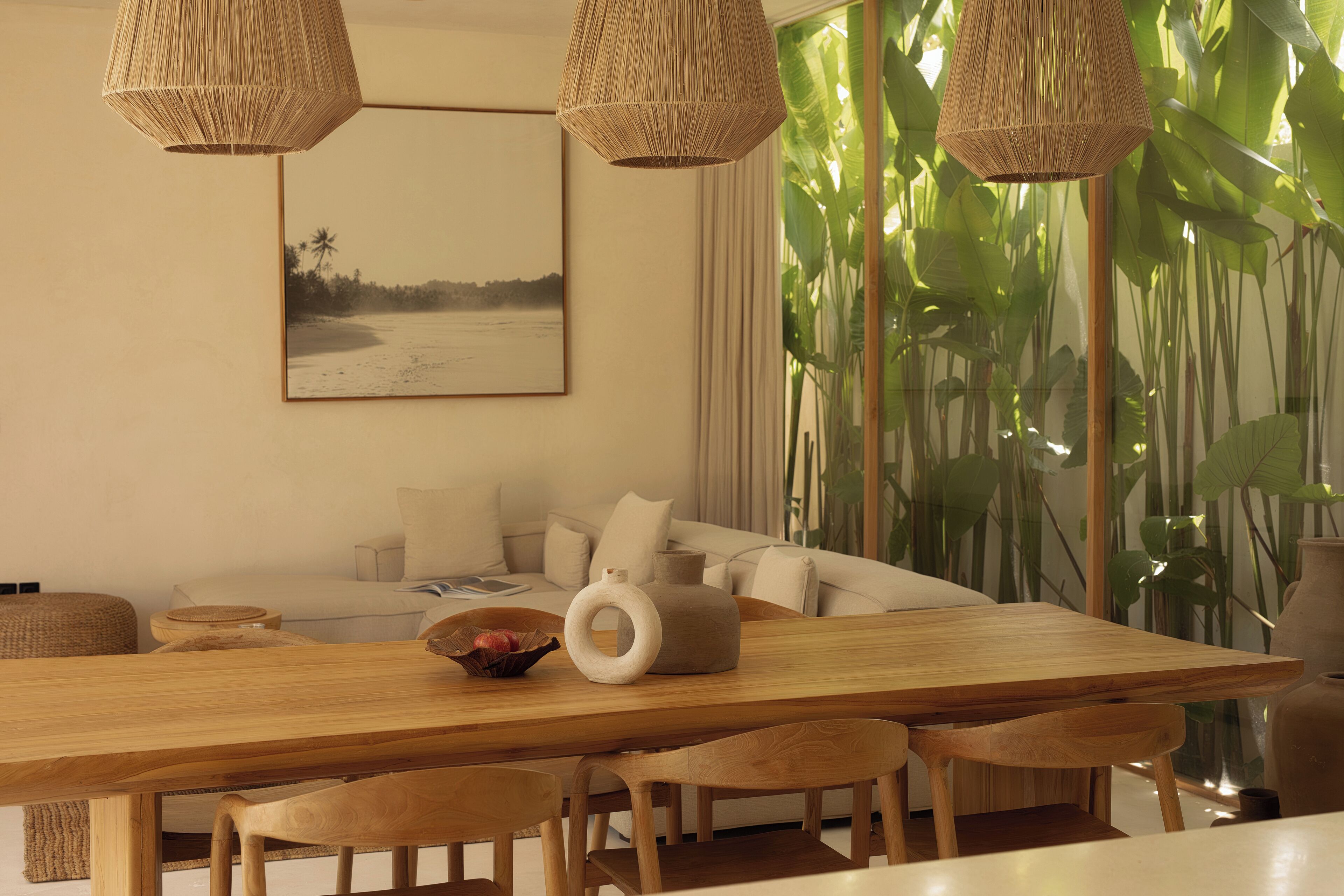 Un comedor con mesa de madera, lámparas colgantes de mimbre y un ventanal con plantas tropicales.