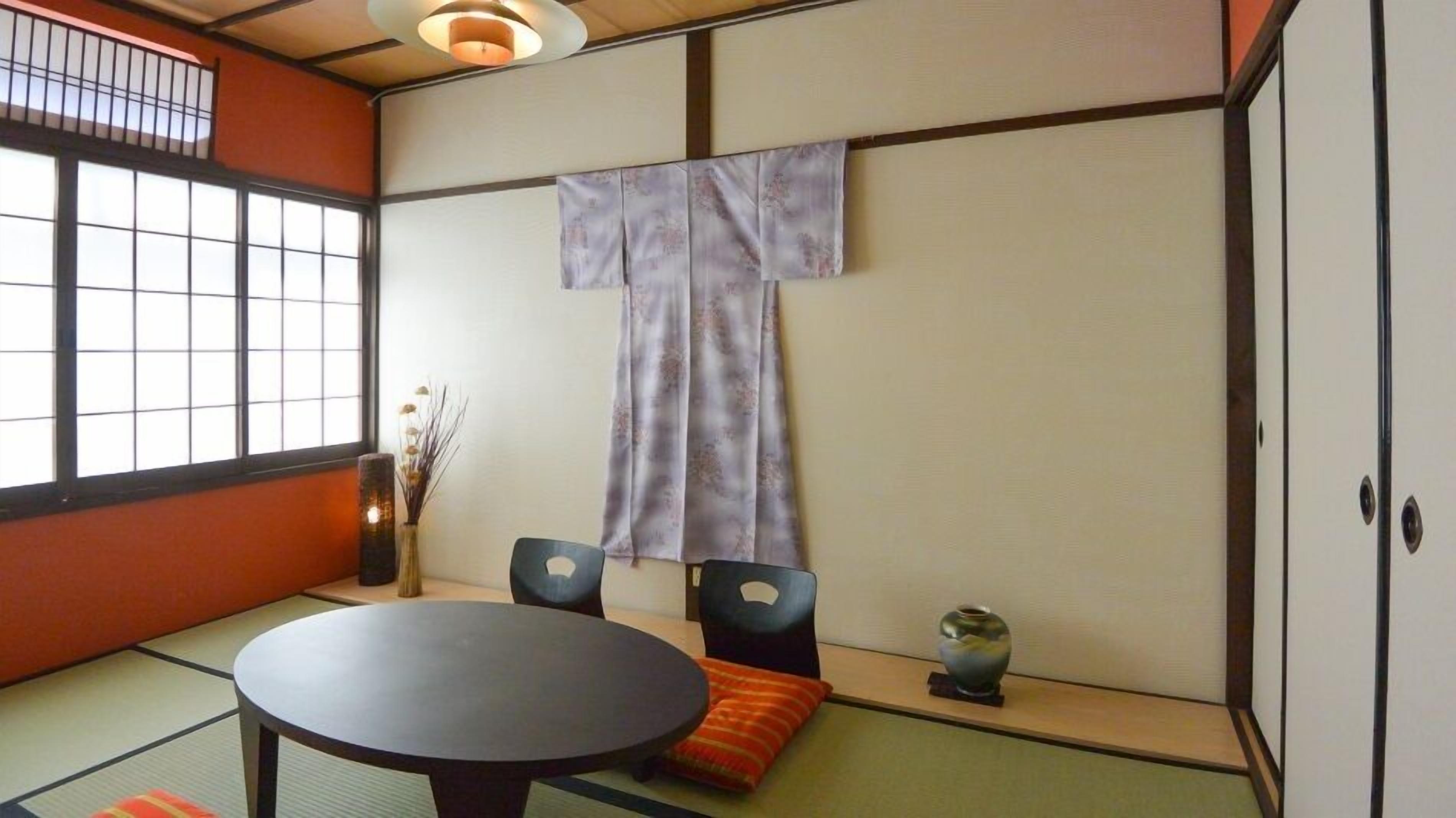 A traditional Japanese room with tatami flooring, a low table, two chairs, a hanging shoji screen, and a small vase on the floor.