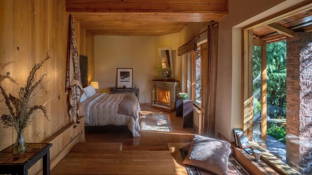 A cozy bedroom with a wooden ceiling, a bed, a fireplace, and a large window with a view of greenery. - Mary Budden Estate Binsar (Kaligari)