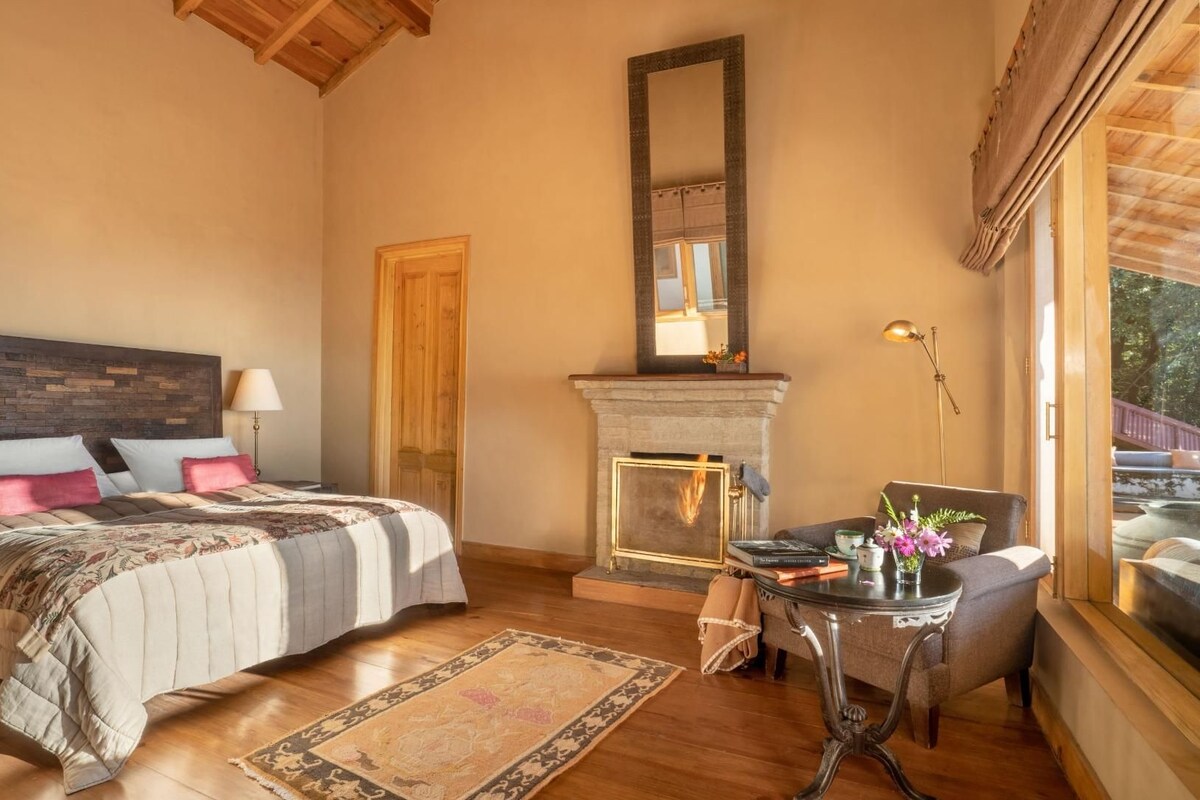 A bedroom with a bed, a fireplace, a table with a vase, and a window with a view.