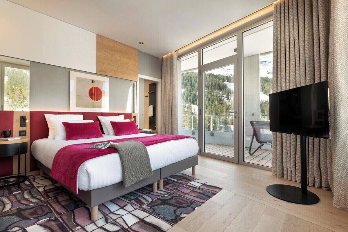 Una habitación de hotel moderna con una cama grande, televisión y balcón con vistas a las montañas.
