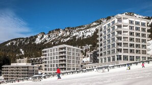 Um hotel moderno com vários andares e varandas, com montanhas cobertas de neve e céu claro ao fundo.
