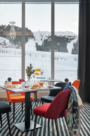 Un comedor con una mesa preparada para dos personas, un ventanal con vistas a una montaña nevada y sillas de colores.
