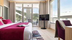 Quarto de hotel com cama, televisão, escrivaninha e vista para montanhas cobertas de neve.