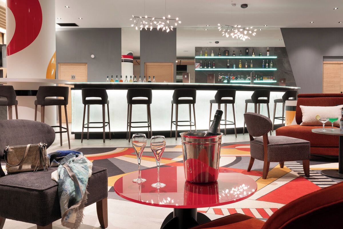 A modern bar area with a bar counter, multiple stools, and a seating area with a red round table.