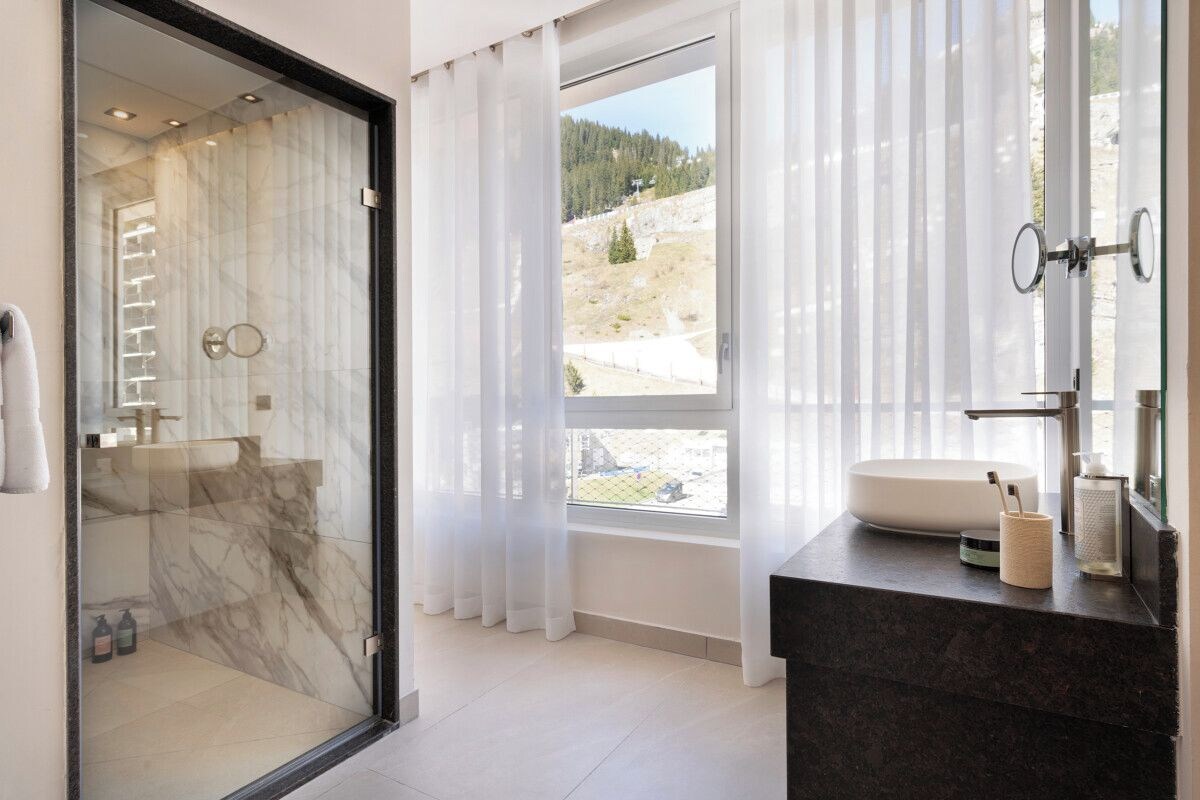 A modern bathroom with a glass-enclosed shower, a large window with a view of a mountainous landscape, and a dark marble countertop with a sink and toiletries.