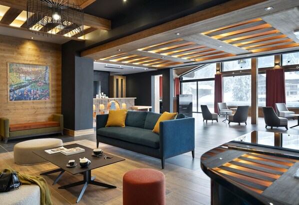 A modern hotel lobby with a wooden ceiling, a large sofa with cushions, a coffee table, and a seating area with chairs and a red ottoman. - MGM Hôtels et Résidences - Hameau De L'ours (Manigod)