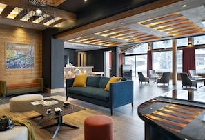 Un moderno vestíbulo de hotel con techo de madera, un sofá grande con cojines, una mesa de centro y una zona de estar con sillas y un reposapiés rojo.