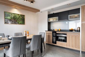 A modern kitchen with wooden cabinets, a built-in oven, and a dining area with grey chairs and a wooden table. - MGM Hôtels et Résidences - Hameau De L'ours (Manigod)