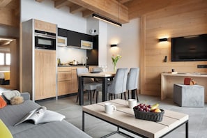 A modern kitchen with built-in appliances, a dining area with a table and chairs, a living area with a sofa, and a TV mounted on the wall. - MGM Hôtels et Résidences - Hameau De L'ours (Manigod)