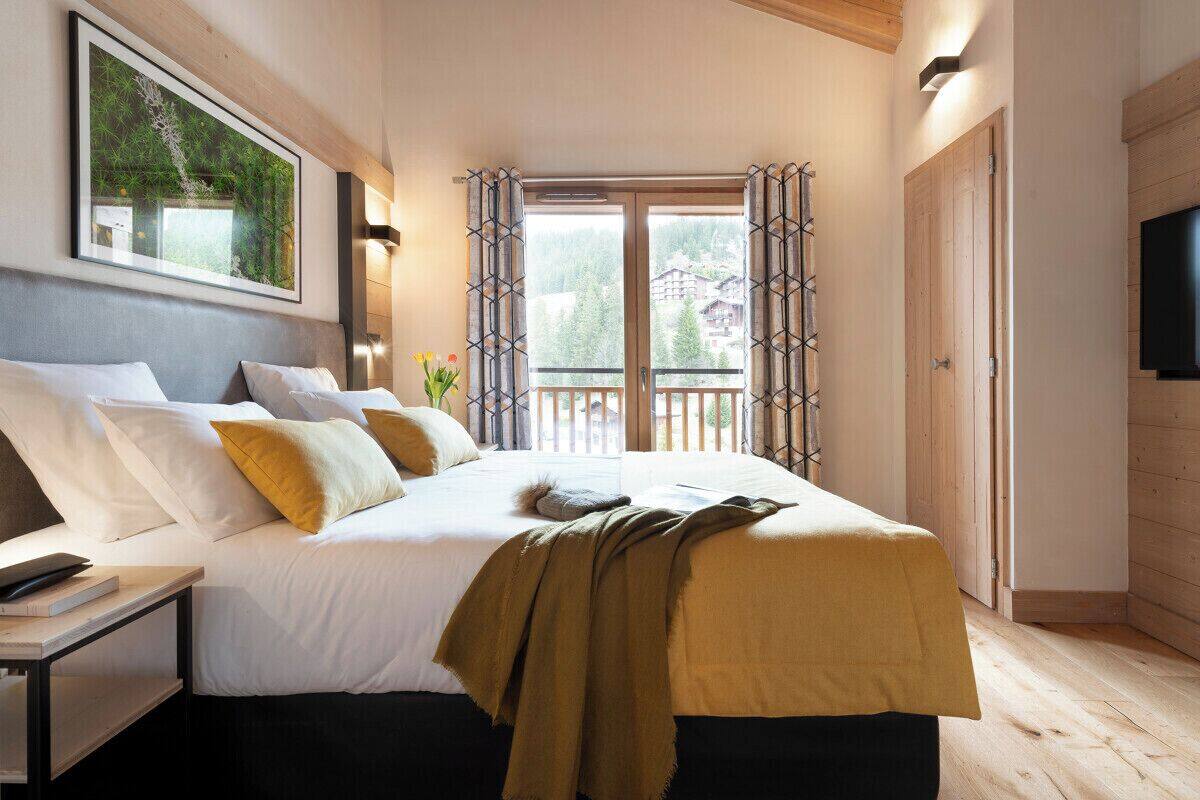 Une chambre avec un grand lit, une table de chevet sur laquelle repose un livre, un tableau accroché au mur et un balcon offrant une vue sur les arbres et les bâtiments.