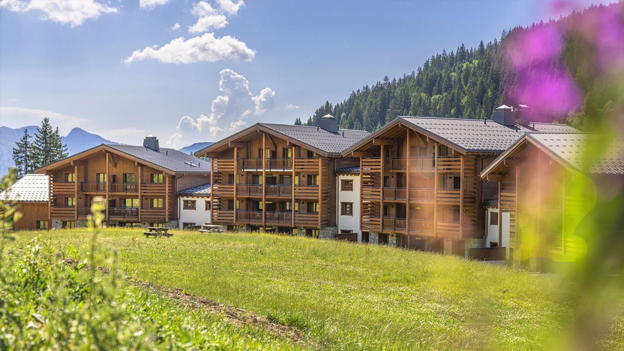 A row of wooden cabins with balconies, set in a grassy field with mountains in the background.