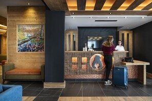A reception area with a wooden reception desk, a large TV screen, and a painting on the wall. - MGM Hôtels et Résidences - Hameau De L'ours (Manigod)