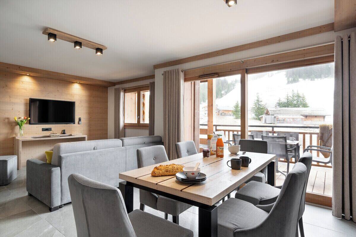 Un salon moderne avec un canapé, une table à manger et des chaises. Un téléviseur à écran plat est fixé au mur, et il y a une fenêtre avec des rideaux.