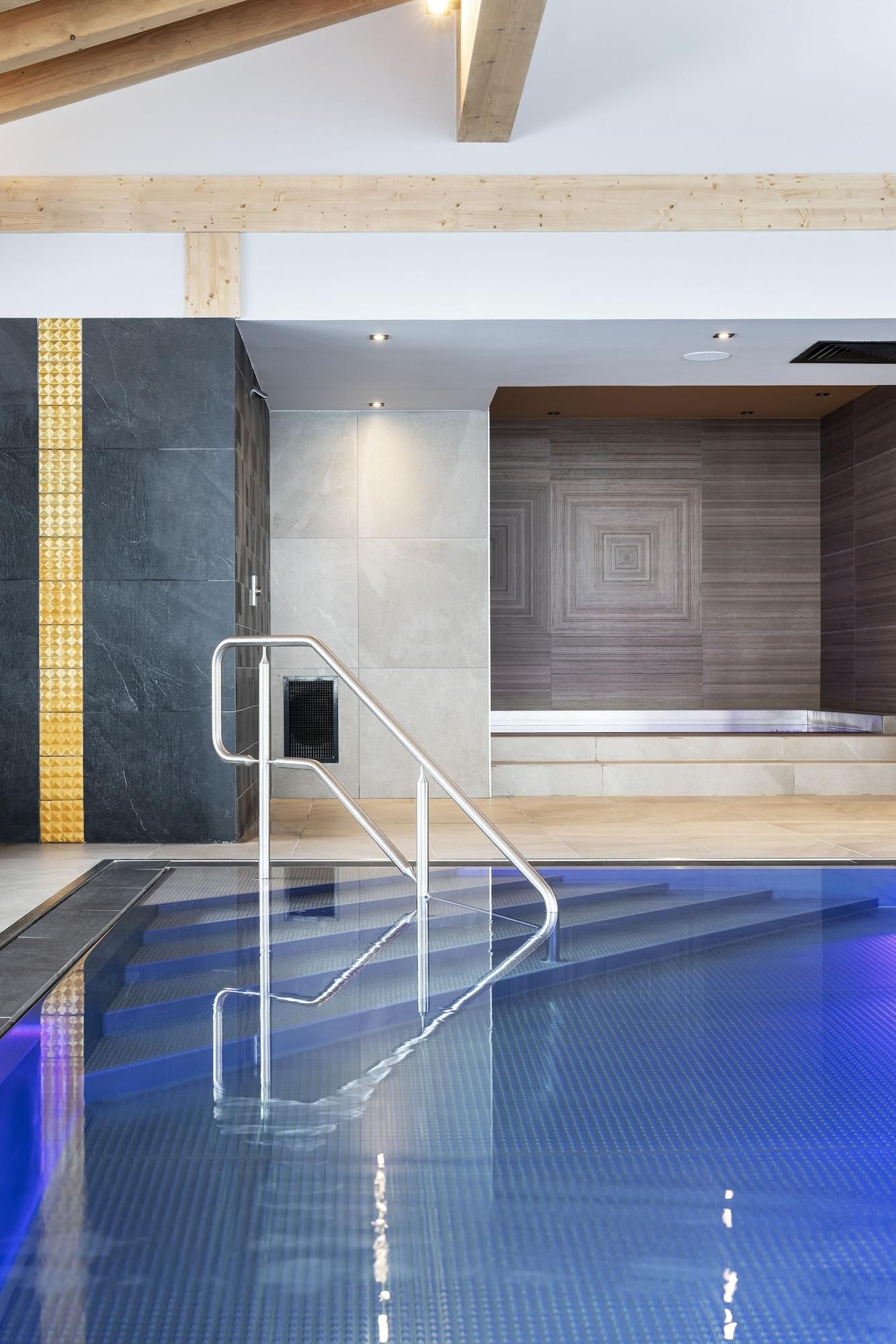 A modern indoor swimming pool with clear glass sides, a metal handrail, and a geometric patterned wall in the background.