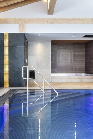 A modern indoor swimming pool with clear glass sides, a metal handrail, and a geometric patterned wall in the background. - MGM Hôtels et Résidences - Hameau De L'ours (Manigod)