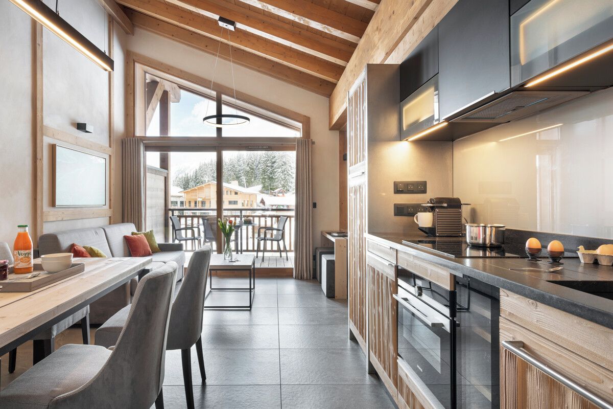 A modern kitchen with a large island, wooden ceiling beams, and a view to the outside.