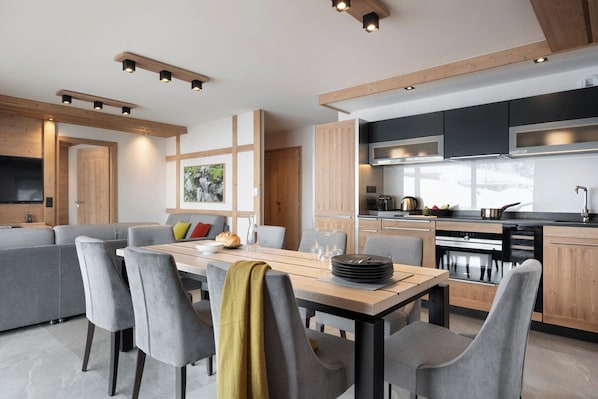 A modern kitchen with built-in appliances, a dining area with grey upholstered chairs, and a wooden dining table. - MGM Hôtels et Résidences - Hameau De L'ours (Manigod)