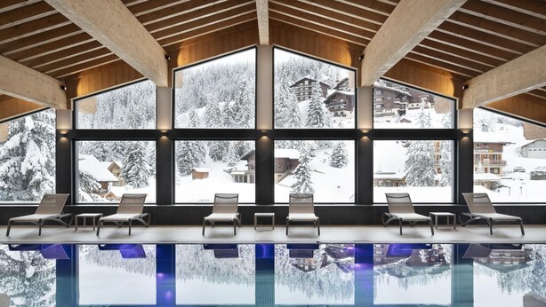 Ein moderner, überdachter Swimmingpool mit großen Fenstern, die einen Blick auf eine verschneite Berglandschaft bieten.