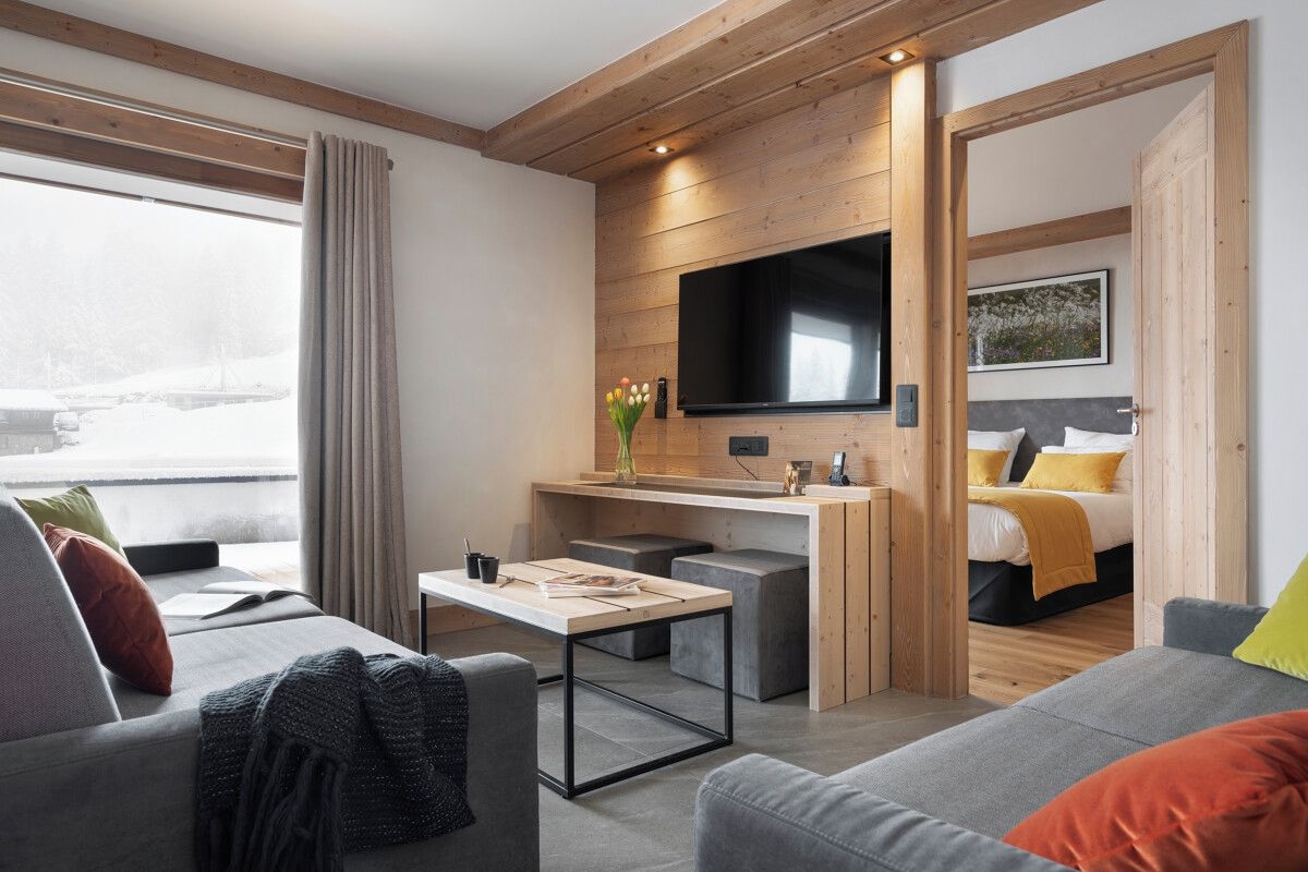 A modern hotel room with a wooden ceiling, a flat-screen TV mounted on the wall, a small kitchenette, a sofa, and a bed with yellow pillows.