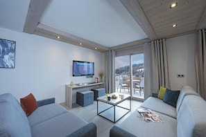 A modern living room with a large flat-screen TV, grey sofas, a coffee table, and a balcony with a view. - MGM Hôtels et Résidences - Hameau De L'ours (Manigod)
