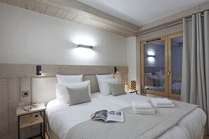 A bedroom with a wooden ceiling, a bed with white bedding, a bedside table with a lamp, and a window with curtains. - MGM Hôtels et Résidences - Hameau De L'ours (Manigod)