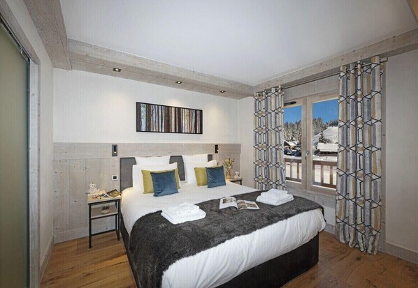 A modern bedroom with a large bed, a nightstand, a wall-mounted artwork, and a view of a snowy landscape through a window with patterned curtains. - MGM Hôtels et Résidences - Hameau De L'ours (Manigod)