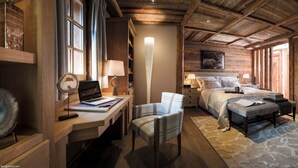 Una stanza accogliente e ben illuminata con soffitto in legno, un letto con una grande finestra, una scrivania con un portatile, una sedia e un tavolino con una lampada.