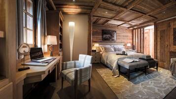 A cozy, well-lit room with a wooden ceiling, a bed with a large window, a desk with a laptop, a chair, and a small table with a lamp.