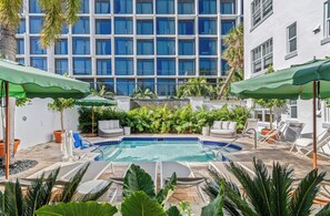 A pool area with lounge chairs and umbrellas, surrounded by lush greenery and a building with large windows in the background. - Aloha Fridays Miami Beach (Bal Harbour)