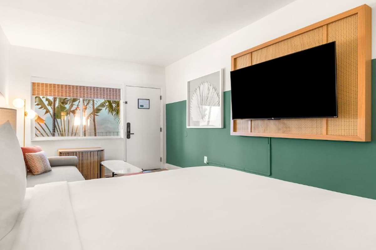 A hotel room with a bed, a desk, a chair, a TV mounted on the wall, and a window with a view of palm trees.