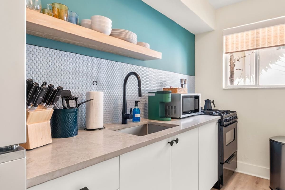 A modern kitchen with white cabinets, a teal accent wall, a stainless steel sink, and a window with a view of trees.