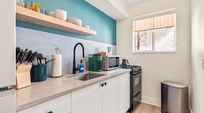 A modern kitchen with white cabinets, a teal accent wall, a stainless steel sink, and a window with a view of trees. - Aloha Fridays Miami Beach (Bal Harbour)