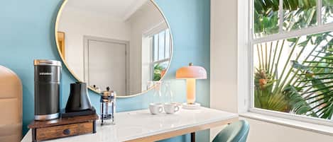 A modern room with a round mirror, a coffee maker, a lamp, and a window with a view of palm trees.