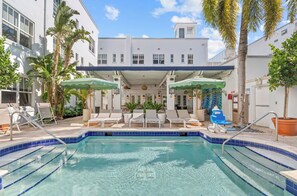 A pool area with lounge chairs and umbrellas, surrounded by palm trees and a building with multiple windows. - Aloha Fridays Miami Beach (Bal Harbour)