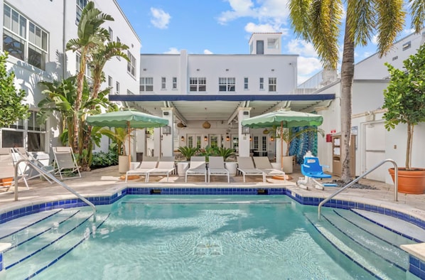 A pool area with lounge chairs and umbrellas, surrounded by palm trees and a building with multiple windows. - Aloha Fridays Miami Beach (Bal Harbour)