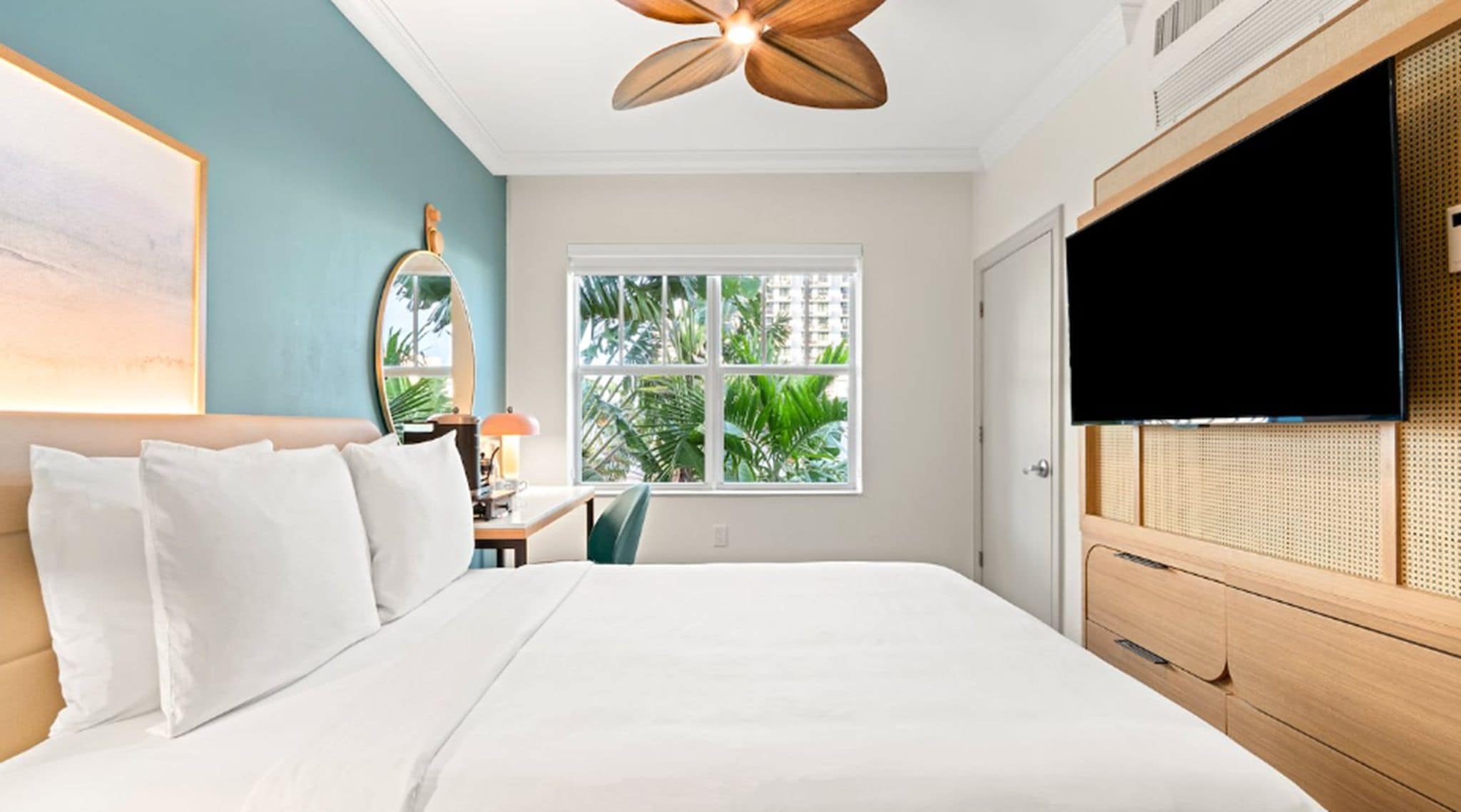 A hotel room with a bed, a ceiling fan, a TV, a window with a view of palm trees, and a wooden dresser.