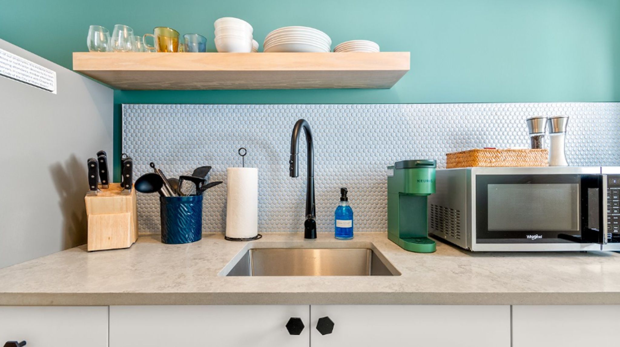 Cozinha com pia de aço inoxidável, micro-ondas e máquina de café. Há utensílios em um recipiente azul, um porta-toalhas de papel branco e uma prateleira de madeira com pratos e copos.
