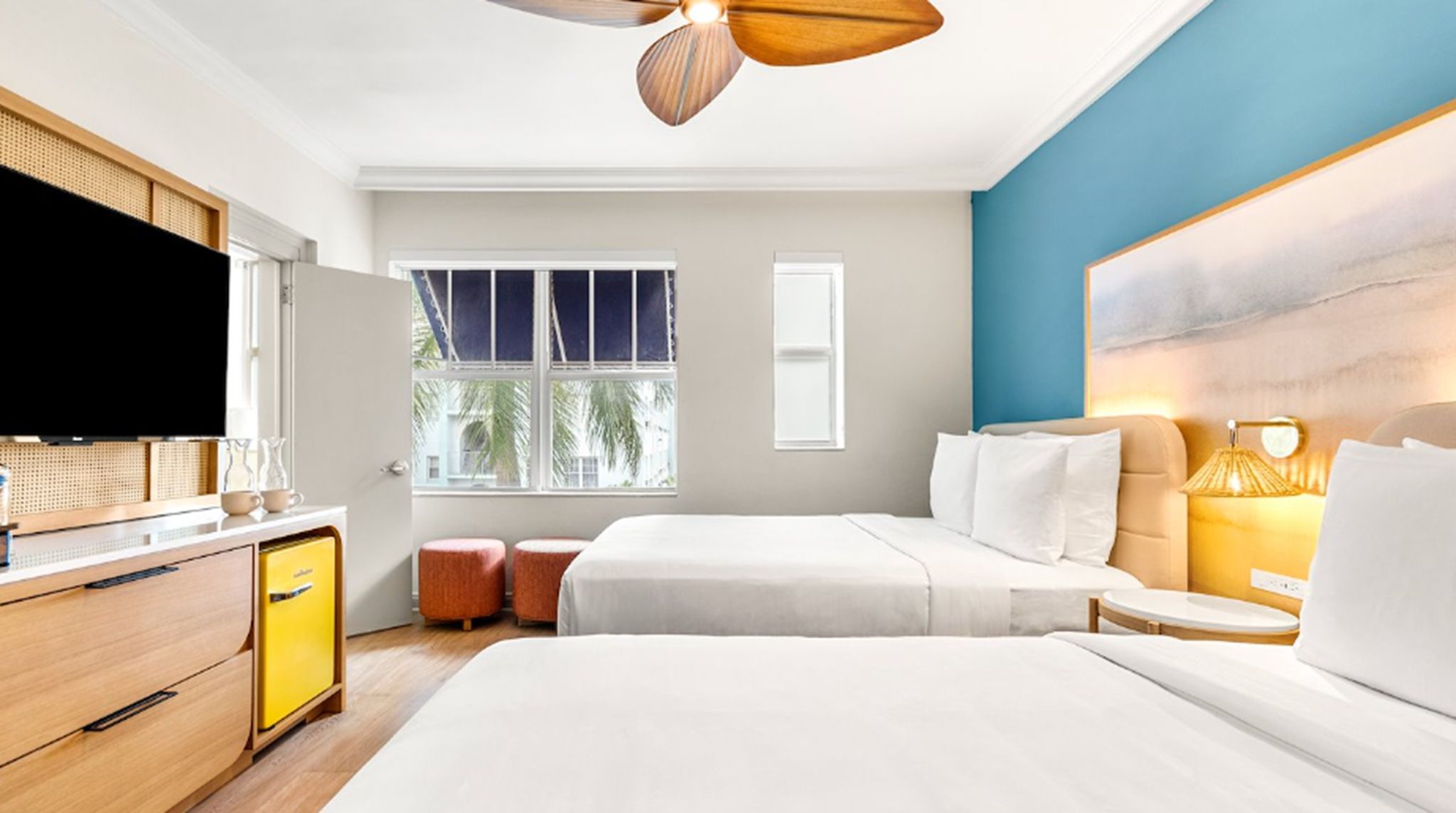 A hotel room with a bed, bedside table, lamp, TV, wardrobe, and a window with a view of palm trees.