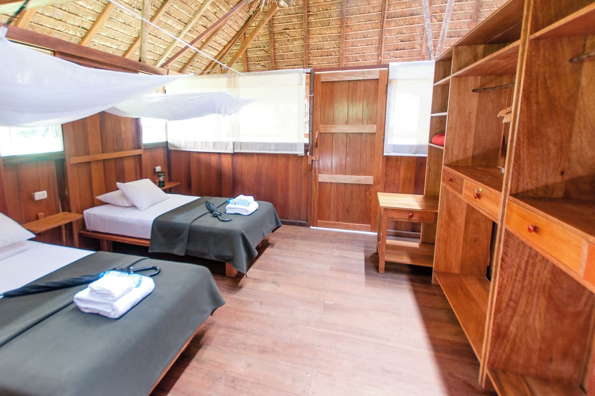 A room with two beds, wooden walls, a wooden desk, and a thatched roof.