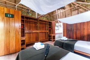 A room with wooden walls and a thatched roof, featuring two beds with towels and an umbrella, a wooden wardrobe, and a number sign indicating room 13. - Amazon Field Station Byinkaterra (Tambopata)